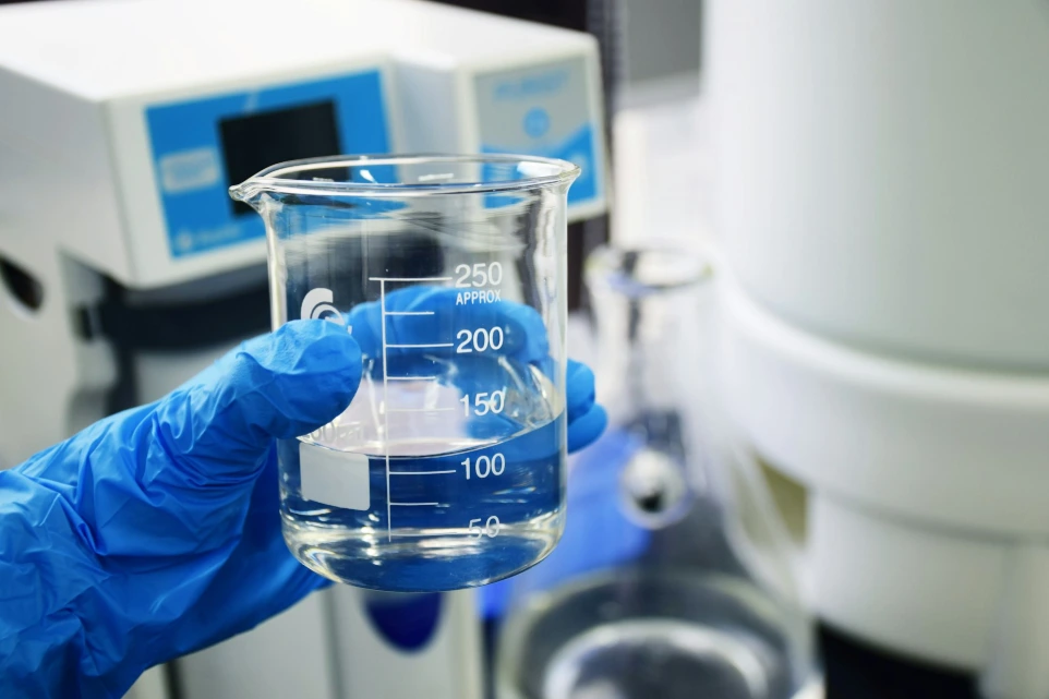 A gloved hand holding a glass beaker filled with clear liquid in a laboratory huasun